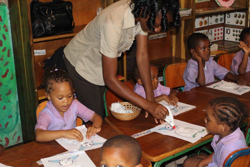 A teacher with her students doing an activity, while she helps one student add glue to her page.
